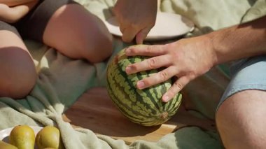 Baba ve kızı pikniğin tadını çıkarıyor, adam taze karpuzu dilimlere ayırıyor. Babam aile yemeği için sulu kırmızı meyveleri dikkatlice dilimliyor. Ellerin ustalıkla olgun karpuz keserken kızın yanında oturuyor.