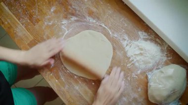 Elleri ahşap yüzeyde yuvarlanan hamurun yanında merdane ve un var. Un kaplı masada yuvarlanan aletle çalışırken parmaklar ince hamur çarşaflarını şekillendiriyor. Eller hamuru düzleştiriyor.