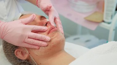 Profesyonel kadın kozmetik uzmanı müşterinin yüzüne temizlik ürünüyle masaj yapıyor. Dişi, cilt bakımı sırasında nazik hareketler uyguluyor. Bayan yüz bakımını titizlikle yapıyor.