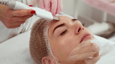 Güzellik kliniğinde yüz bakımı sırasında ultrasonik cilt fırçası kullanan bir profesyonel. Yumuşak bir deri yüzme ve ileri teknoloji ile derin bir temizlik. İstemci alınırken dinleniyor