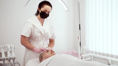 Kozmetolog güzellik uzmanı kozmetoloji kliniğinde tedavi prosedürlerinden önce yüzünü temizliyor. Maske takan kadın uzman ve eldivenler nazikçe deriyi terapi için hazırlıyor. Bayan Güzellik Uzmanı