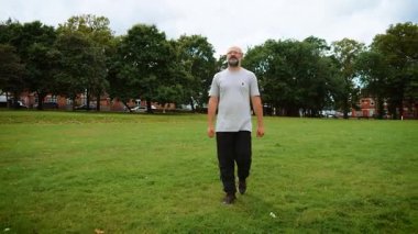 Adam sakin bir tavırla parkta yürüyor. Yetişkin adam kendini rahat ve odaklanmış hissediyor. Orta yaşlı, açık tişörtlü, siyah pantolonlu ve sağlam ayakkabılı bir adam. Sakallı adam açık otlakta geziniyor.