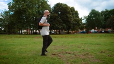 Sırt çantası taşıyan bir adam parkta yürüyor. Yetişkin adam sakin ve rahat görünüyor. Orta yaşlı, açık tişörtlü, siyah pantolonlu ve sıradan ayakkabılı bir adam. Sakallı adam otlakta sırt çantasıyla dolaşıyor.