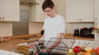 Genç adam mutfak lavabosunda ahşap kesme tahtasını duruluyor, temizliğe odaklanmış. Erkek yıkama tahtası akan suyun altında, işine odaklanıyor. Adam mutfak malzemelerini suyla temizliyor.