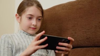 Kanepede otururken akıllı telefonda oynayan bir kız. Çocuk mobil aktivite sırasında ekrana odaklanıyor. Çocuk boş vakitlerinde telefonu kullanıyor. Aygıt üzerinde dijital etkileşime giren kişi