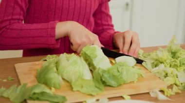 Tahtada marul kesen eller. Parmaklar taze yeşillikleri bıçakla kesiyor. Yemek yapmak için marul doğrayan ellerin yakın çekimi.