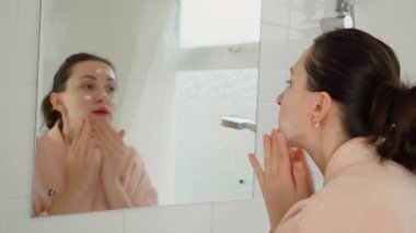 Aynanın önünde yüzüne krem süren bir kadın. Yansımaya bakarken cilt bakımı yapan bir kadın. Sıvı almak için cilde masaj yapan kadın kremi. Yüzü nemlendirici ile besleyen kişi