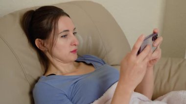 Kadın serin odada akıllı telefon kullanarak battaniyeye sarılı olarak kanepede yatıyor. Küresel gaz krizi nedeniyle soğuk hava şartlarıyla karşı karşıya kalan bir kadın. Donmaya dayanırken meşgul olmaya çalışan bir kadın.