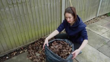 Tahta çitlerin yanındaki çöp poşetine yaprakları koyan kadın. Kuru yaprakları toplayıp poşetleyerek dışarıyı temizleyen kadın. Sonbahar enkazı düzenleyerek teras alanını düzenleyen bir ev hanımı. Hanımefendi.