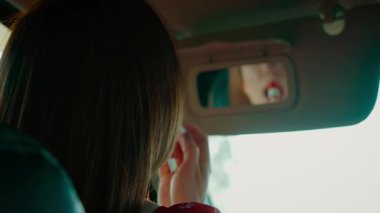 Araba güneşliği aynasına bakarken ruj süren bir kadın. Yansıma, elleri makyajı ayarlarken odak noktasında parlak kırmızı dudaklar gösteriyor. Küçük bir ayna kullanarak aracın içindeki kadına dokunuyor.