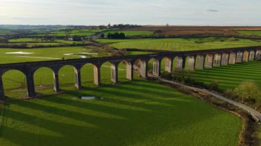 Yumuşak güneş ışığı altında yeşil tarlaların üzerine uzanan tuğla viyadük. Gün batımında parlayan manzaralı viyadük. Tarihi tren köprüsü kırsal alanda altın ışıkla kavis çiziyor