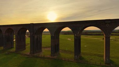 Günbatımında yeşil tarlaların üzerinde yükselen tarihi demiryolu viyaolkü. Demiryolu köprüsü alacakaranlığın sıcak altın ışığıyla aydınlandı. Antik tuğla viyadük zarif bir şekilde akşam ışığı altında manzaranın üzerine eğilmiş.
