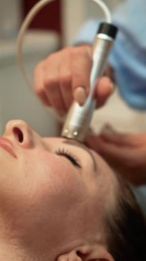 Alnında mikro akım terapisi uygulayan profesyonel cilt bakımı, dikey yönelim. Güzellik uzmanı yüz kaslarını uyarmak için nazik elektrik darbeleri kullanıyor. Deriyi geliştiren estetik uzmanı