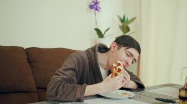 Bornoz giymiş bir adam masada oturmuş sosisli yiyor. Sıcak ev ortamında bir erkek fast food atıştırıyor. Adam içeride sosisli sandviç yiyor.