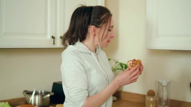 Modern beyaz mutfakta sosisli yiyen bir kadın. Çağdaş yemek alanında ev yapımı atıştırmalıklardan hoşlanan bir kadın. Hanımefendinin yeni hazırlanmış sandviçi şık mutfak alanında tadıyor.