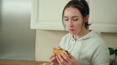 Modern beyaz mutfakta sosisli yiyen bir kadın. Çağdaş yemek alanında taze yapılmış sandviçin tadını çıkaran bir kadın. Şık mutfak alanında ev yapımı aperatifleri tadıyor.