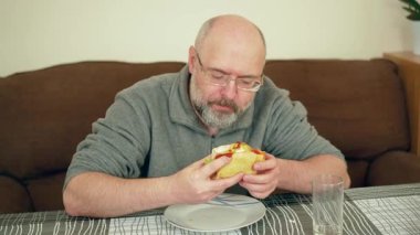 Yemek masasında içi doldurulmuş sandviç yiyen bir adam. Erkek, soslu ve soslu çıtır ekmeği ısırıyor. Adam rahat bir evde ev yemeği yiyor. İçerideki pis ve lezzetli atıştırmalıkların tadını çıkaran kişi.