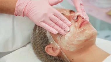 Eldiven takan kozmetolog yumuşak hareketlerle yüz temizliği yapıyor. Tedavi sırasında profesyonel cilt bakımı ürünleri eşit düzeyde. Nazikçe ve titizlikle pürüzsüz bir uygulama sağlayan uzman.