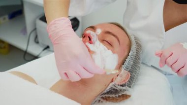 Kadın rahat bir şekilde uzanıyor, gözleri kapalı, profesyonel kozmetik uzmanı beyaz kremi ahşap aplikatörle yüzüne sürüyor. Dişi, krem eşit olarak yayılırken rahatlatıcı cilt bakımı yapmaktan hoşlanıyor. Hanımefendi.