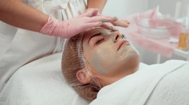 Kadın yüz maskesi uygulaması alıyor, cilt bakımı sırasında sakince yatıyor. Güzellik uzmanı nazikçe yüzünü maskeyle kapatıyor. Hasta kozmetoloji kliniğinde dinleniyor.