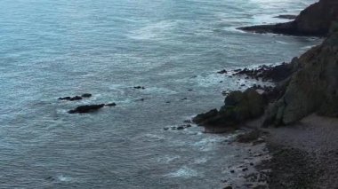 Engebeli kıyı şeridi boyunca kayaların üzerinde okyanus dalgaları kırılıyor. Deniz sörfü kıyıya yakın pürüzlü taş oluşumlarına çarpıyor. Su bulutlu gökyüzünün altında kayalık sahile vuruyor. Dalgalar dalgalı kıyıları vuruyor.