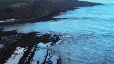 Kıyı köyü kayalık kıyı şeridinde akşam ışığını yansıtan dalgalarla. Küçük bir sahil kasabası dramatik uçurum ve okyanusun kenarında dinleniyor. Güneş batarken engebeli kıyı şeridi boyunca uzanan sessiz evler. Rıhtım