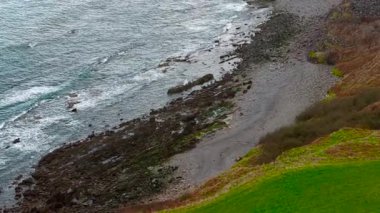 Okyanus dalgaları çimenli yamaçtaki kayalık plaja çarpar. Kıyı şeridine vuran deniz sörfü taşlarla ve dallarla dağılmış. Gel-git suları yeşil yamaçların yanındaki engebeli kıyılara vurur. Yuvarlanan dalgalar kırılıyor