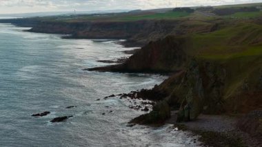 Uzun kayalık kıyı şeridi okyanus boyunca kayalıklar ve dalgalarla uzanıyor. Bulutlu gökyüzünün altında dalgalanan engebeli deniz kıyısı. Sivri kıyı şeridi uzak burunların ötesinde denizle buluşuyor. Çarpıcı sörf vuruşu