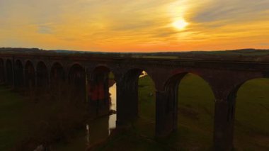 Gün batımında huzurlu kırsal kesimde uzanan demiryolu viyameti. Akşam gökyüzü sıcak renklerde viyadük boyuyor. Altın alacakaranlık altında yemyeşil vadi boyunca uzanan devasa tuğla köprü.