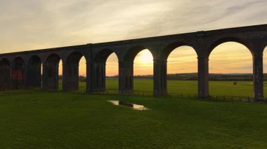 Muhteşem tuğla viyadük gün batımında açık yeşil tarlalar üzerinde kemer. Tarihi viyadük ile huzurlu günbatımı sahnesi. Altın gökyüzü altında engin kırlara yayılan demiryolu köprüsü