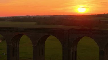 Büyük tuğla viyadük yeşil tarlalar üzerinde kavis alevli günbatımı gökyüzü altında. Viyadük, alacakaranlık gökyüzü altında doğayla uyum içinde. Huzur içinde uzanan demiryolu köprüsü.