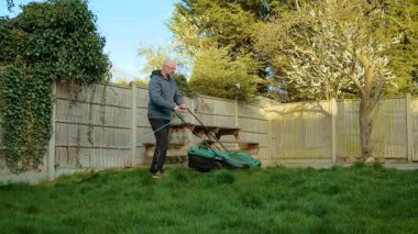 Bahçede elektrikli çim biçme makinesiyle çim biçen bir adam. Bahçe çimlerini kordonlu çim biçme makinesiyle kesen bir erkek. Ahşap çitlerin yanında yeşil çim biçme makinesiyle kalın çimler kesen adam.
