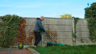 Arka bahçede elektrikli çim biçme makinesiyle çim biçmek. Ahşap çitlerin yanında çim biçme makinesi kullanarak çimleri kesmek. Dışarıdaki elektrikli bahçe aletleriyle aşırı gelişmiş çimenleri buduyorum.