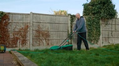 Arka bahçedeki çimenleri elektrikli çim biçme makinesiyle buduyor. Erkek, çimleri kordonla biçiyor. Bahçede yeşil çim biçme makinesiyle kalın çim biçen bir adam var.