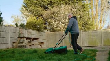 Adam arka bahçede elektrikli çim biçme makinesiyle uzun çimleri kesiyor. Ahşap çitlerin yakınındaki çimleri biçmek için yeşil çim biçme makinesi kullanan bir erkek. Dışarıdaki çimlerin bakımı için kablosuz bahçe aracı