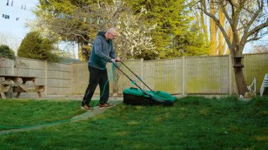 Bahçede elektrikli çim biçme makinesiyle çim biçen bir adam. Kordonlu çit kullanarak çitlerin yanındaki erkek kırpma tesisi. Dışarıdaki çimenlik alanı elektrikli bahçe aletleriyle kesen adam.