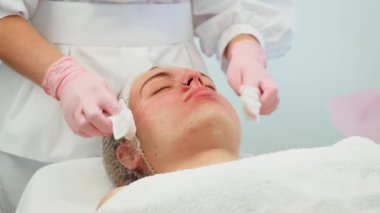 Kozmetik uzmanı, hasta yüzüne pamuk pedi uygulayarak terapi yapıyor. Profesyonel cilt bakımını hassasiyetle ve özenle yapıyor. Uzman, bu süreçte kapsamlı temizlik sağlıyor.