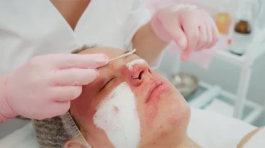 Rahatlamış bir kadın, göze çarpan bir burun halkası, kozmetik uzmanı, tahta sopayla yüzüne krem sürüyor. Cilt bakımına maruz kalan bir kadın, profesyonel cilt ürünüdür. Bayan yüz bakımını seviyor.