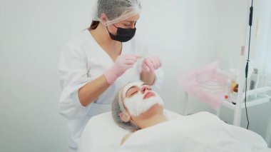 Tıbbi klinikte profesyonel cilt bakımı sırasında rejenerasyon kremi süren bir kadın. Dişi, cilt onarımı ve su kaybını arttırmak için cilt yenileme üzerinde dikkatle çalışıyor. Rahat uzanan kız.