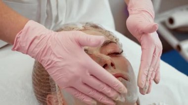 Yenilenme tedavisi gören kadın seans sırasında yüz maskesi ile rahatça yatıyor. Güzellik uzmanı cilt bakımını geliştirmek için yüze nazikçe masaj yapıyor. Hasta kozmetolojide rahatlatıcı terapinin keyfini çıkarıyor