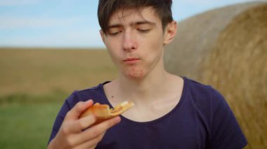 Sahada pizza yiyen genç bir çocuk balya arka planda, sağlıksız fast food. Genç bir erkek dışarıda pizzanın tadını çıkarıyor. Genç çocuk huzurlu kırsal kesimde fast food 'un tadını çıkarıyor.