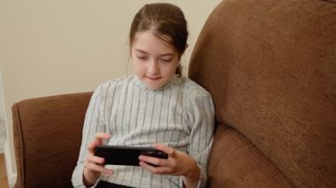 Kanepede otururken akıllı telefona bakan bir kız. Çocuk aktivite sırasında mobil cihaza odaklanmış. Çocuk eğlence olsun diye telefonla meşgul oluyor. Boş zamanlarında ekranla etkileşim halinde olan kişi