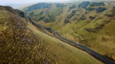 Dik yeşil vadide virajlı yolda giden arabalar. Araçlar İngiltere 'nin Peak District bölgesindeki manzaralı dağ yolunda ilerliyor. Engebeli yamaçlardan geçen virajlı yolun hava görüntüsü. Arabalar resimli kırsal bölgelerde seyahat ediyorlar.