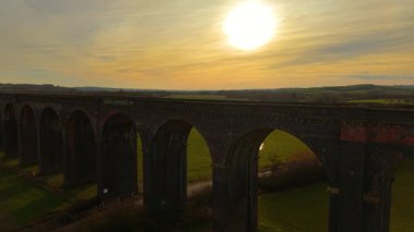 Kırsal kesimde altın gün batımına karşı duran görkemli demiryolu viyadük. Göz kamaştırıcı viyadük, alacakaranlıkta resim gibi manzaraya yayılıyor. Büyük tuğla köprü sıcak alacakaranlık ışıltısıyla yuvarlanan yeşil tarlaların üzerinde