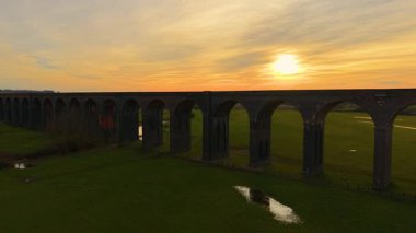 Altın gün batımının altında verimli kırsal bölgelere uzanan uzun tuğlalı viyadük. Iconic demiryolu köprüsü turuncu gökyüzüne gururla karşı duruyor. Geniş tarihi viyadük sıcak akşam ışığında yeşil tarlaları kaplıyor.