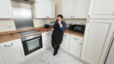 Mutfakta kahve içen bir kadın. Kadın elinde aperatif tutarken sıcak içecekten zevk alıyor. Tezgahın yanında duran bir bayan mola veriyor. İçeride sıcak içecekle rahatlayan biri.