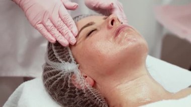 Estetisyen nazikçe alnına masaj yapıyor, hasta dinleniyor. Dermatoloji bürosu çalışanı tam yüz bakımı yapıyor. Cilt bakımı uzmanı rahatlamaya ve rahatlamaya odaklanıyor