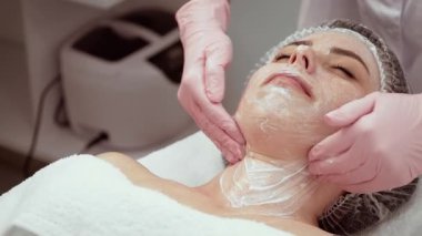 Genç ve güzel bir kadın güzellik prosedürü sırasında sakince kanepede yatıyor. Kozmetik uzmanı boyuna krem sürerek masaj yapıyor, hatta uygulamayı bile garantiliyor. Cilt bakımı uzmanı rahatlatıcı ve gençleştirici tedavi uyguluyor.