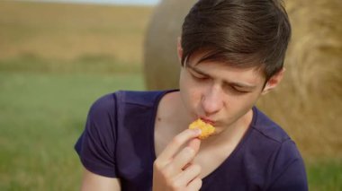 Genç bir delikanlı tavuk nugget yiyor, kızarmış et yiyor, kırsal alanda sağlıksız yiyecekler yiyor. Genç erkek kırsalda çıtır çıtır nuggetların tadını çıkarıyor. Genç çocuk, doğal ortamda kızarmış tavuk ısırığının tadını çıkarıyor.