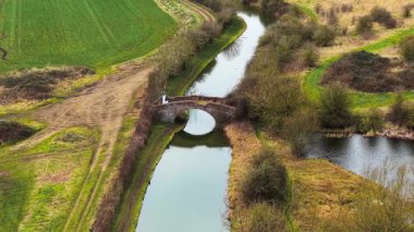 Eski tuğla köprü Birleşik Krallık 'ta tarihi kanalı geçiyor. Sakin su, kırsal alanda kemerli duvarcılık yapısını yansıtıyor. Geniş bankalarla kırsal alanda akan dar su yolu. Kum yolu ile seyrüsefer kanalı boyunca uzanan taş köprü